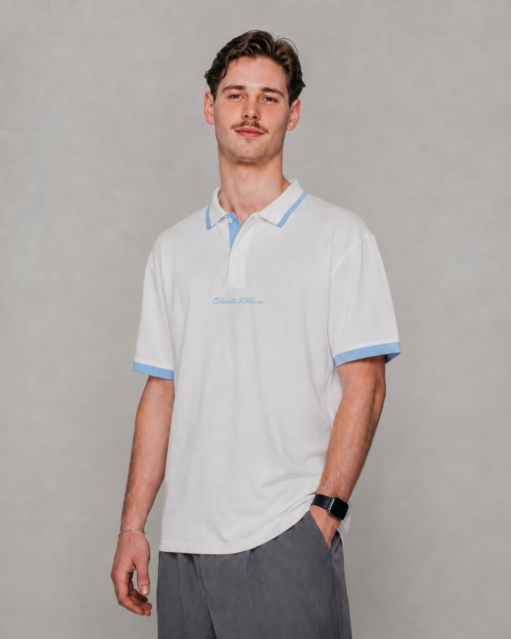 A man wearing a white short-sleeve polo shirt with light blue trim on the collar, sleeve cuffs, and a small light blue embroidered logo on the chest, paired with gray trousers, against a neutral gray background.