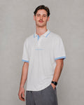 A man wearing a white short-sleeve polo shirt with light blue trim on the collar, sleeve cuffs, and a small light blue embroidered logo on the chest, paired with gray trousers, against a neutral gray background.