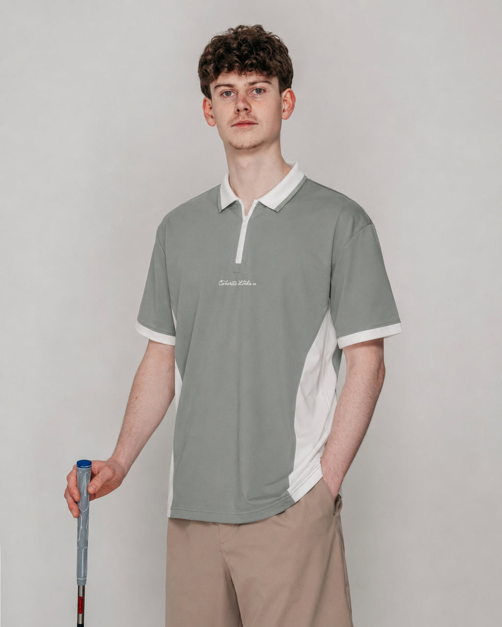 A man modeling a short-sleeve polo shirt in a studio setting. The shirt is sage green with white collar, sleeve cuffs, and side panels, featuring a small white script logo on the left chest.