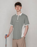 A man modeling a short-sleeve polo shirt in a studio setting. The shirt is sage green with white collar, sleeve cuffs, and side panels, featuring a small white script logo on the left chest.
