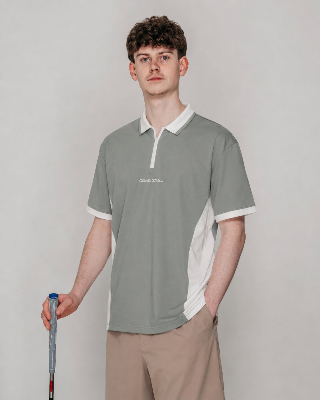 A man modeling a short-sleeve polo shirt in a studio setting. The shirt is sage green with white collar, sleeve cuffs, and side panels, featuring a small white script logo on the left chest.
