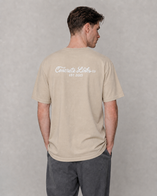 A man viewed from the back wearing a light beige t-shirt with 'Concrete Links co EST 2023' printed in white script on the upper back, paired with dark gray pants, against a textured gray studio background.