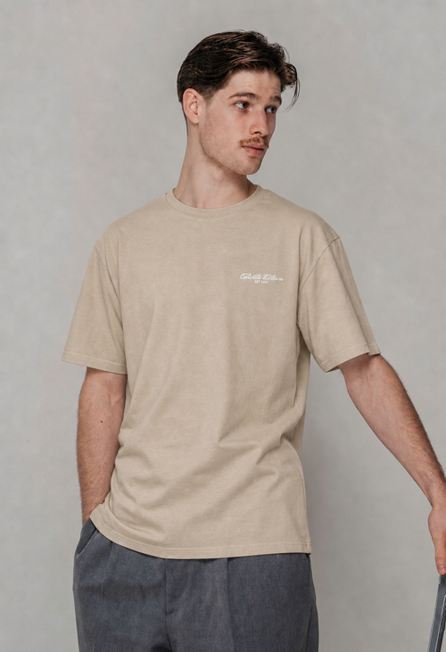 A man wearing a light beige crewneck t-shirt with a small white graphic on the left chest, paired with dark gray pants, standing against a neutral gray background.