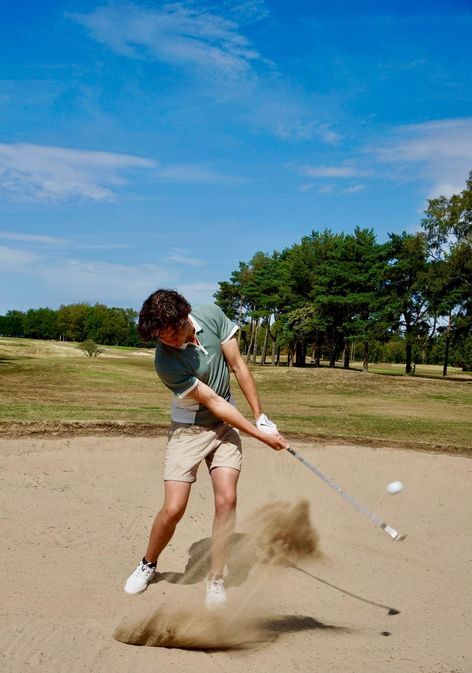 A man in a green polo shirt and khaki shorts swings a golf club from inside a sand bunker on a golf course, kicking up a cloud of sand under a blue sky with trees in the background.