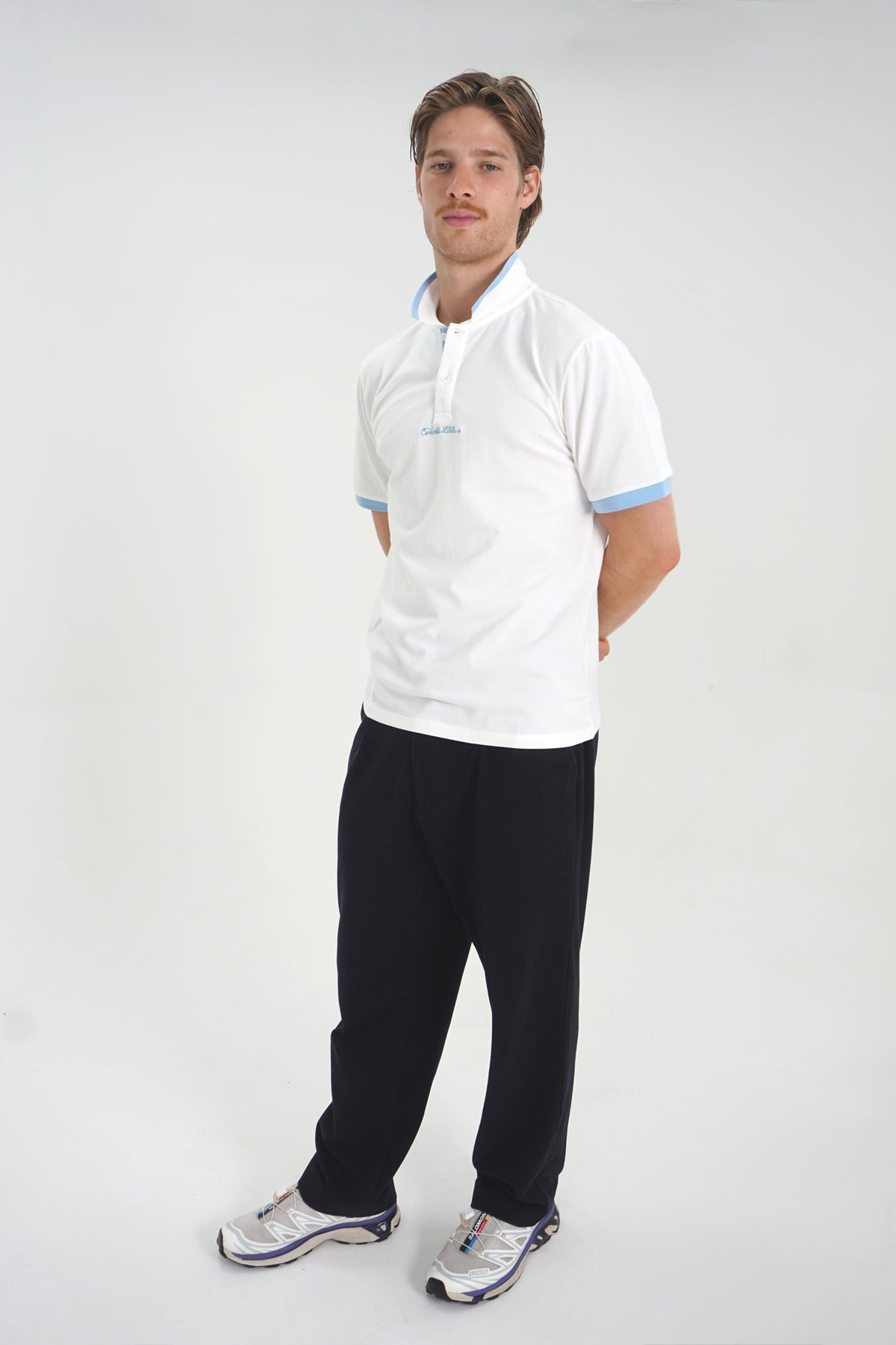A man modeling a white short-sleeve polo shirt with light blue trim on the collar and sleeve cuffs, paired with dark pants and athletic shoes, against a plain light gray studio background.