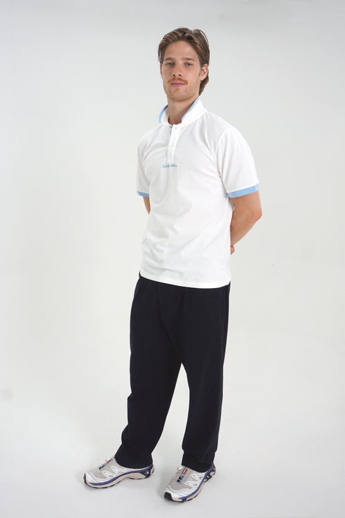 A man modeling a white short-sleeve polo shirt with light blue trim on the collar and sleeve cuffs, paired with dark pants and athletic shoes, against a plain light gray studio background.