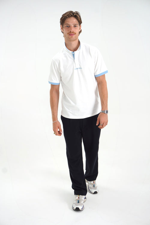 A man modeling a white short-sleeve polo shirt with light blue trim on the collar and sleeve cuffs, paired with black pants and white sneakers, against a plain white studio background.