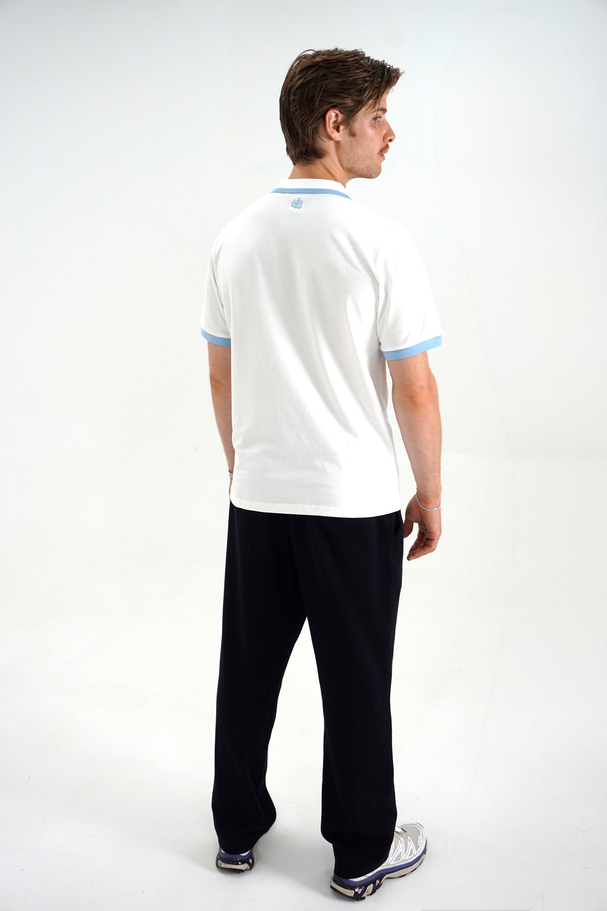A man shown from the back wearing a white short-sleeve polo shirt with light blue trim on the collar and sleeve cuffs, paired with black pants and white sneakers, against a plain white studio background.