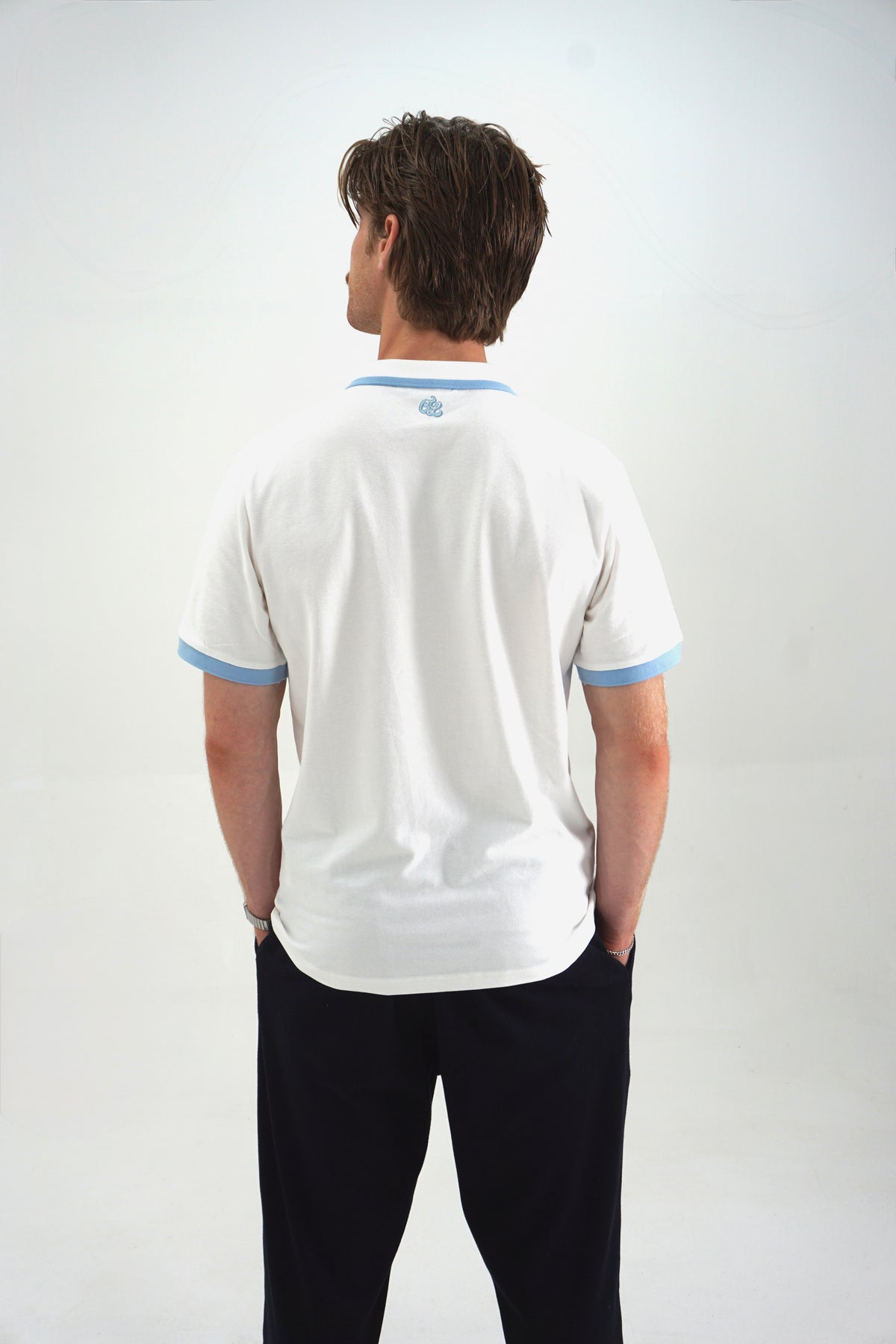 A man viewed from behind wearing a white short-sleeve polo shirt with light blue trim on the collar and sleeve cuffs, paired with dark trousers, against a plain light gray studio background.