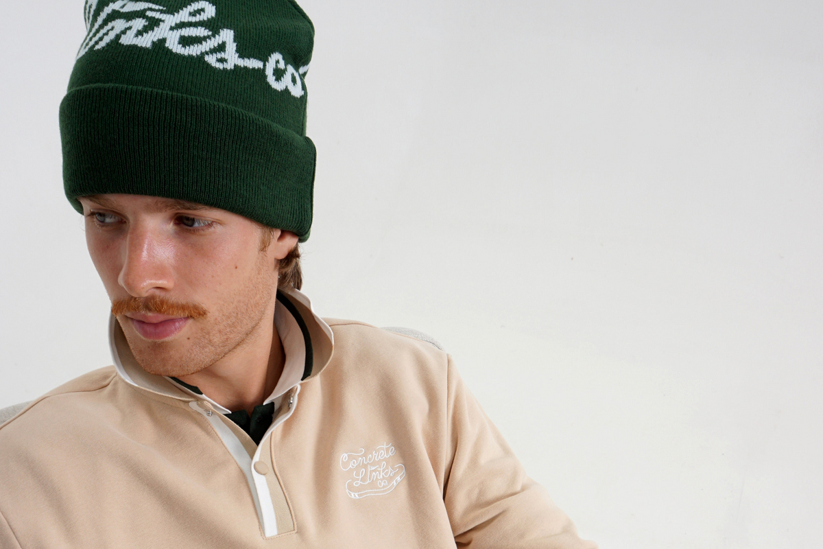 A man wearing a beige quarter-zip pullover sweatshirt, black pants, and a dark green beanie with white script lettering, seated in a chair against a plain light background.