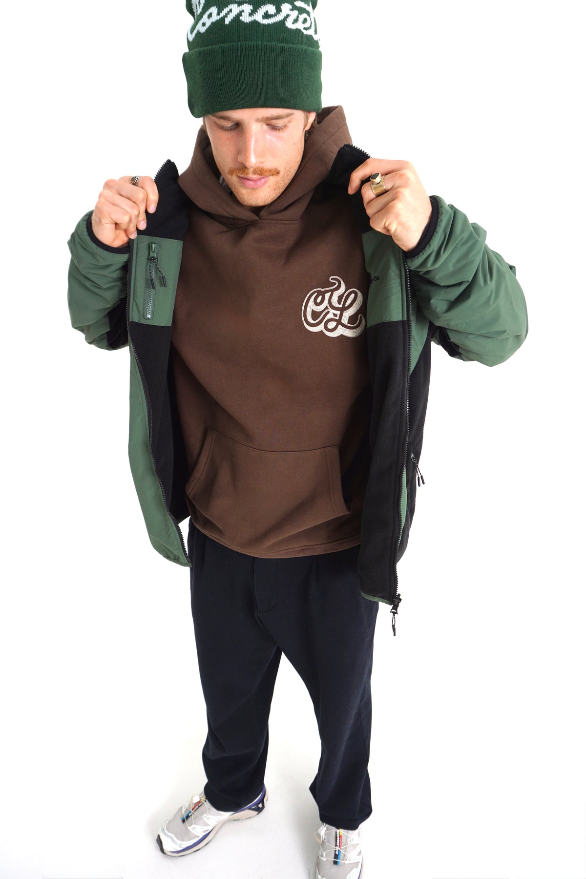 A man wearing a brown hoodie with a white graphic logo, a green and black colorblock jacket, a green beanie with white text, and dark sweatpants, posing against a white studio background.