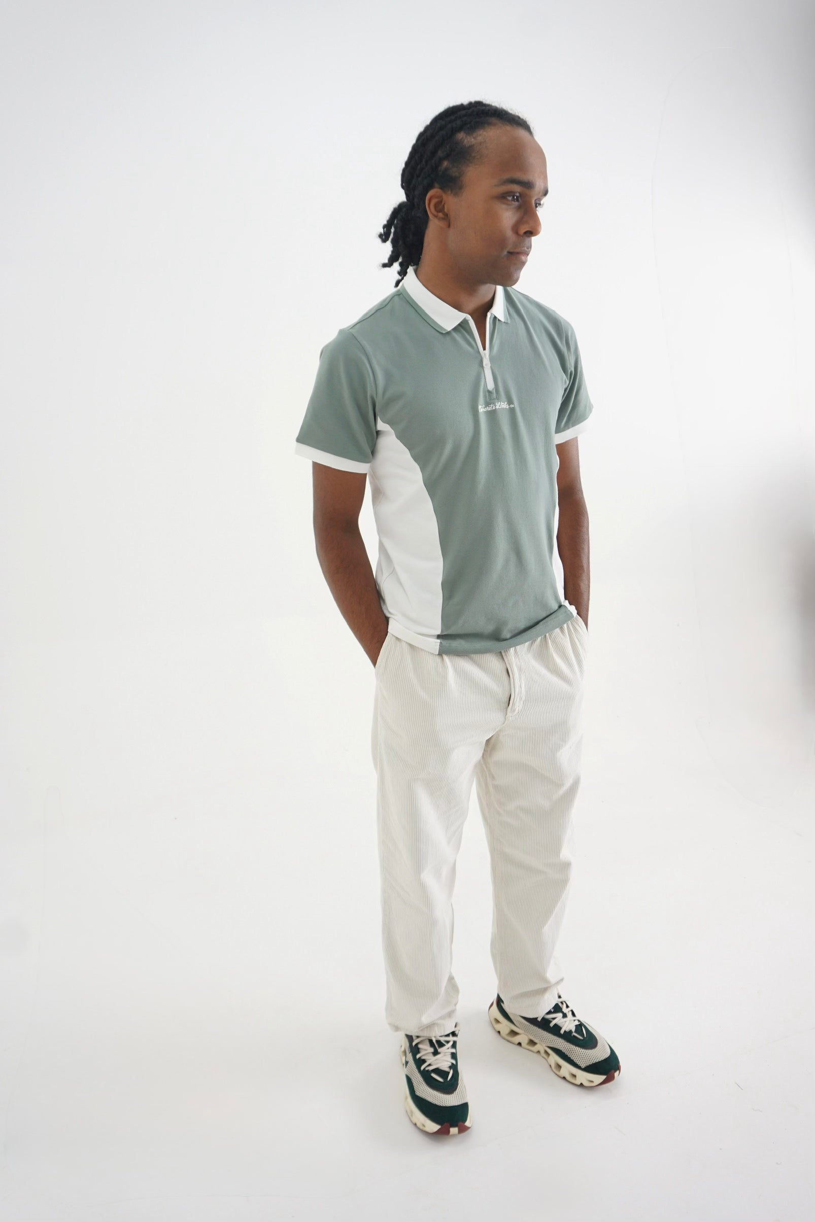A man modeling a short-sleeve polo shirt with a sage green and white color-blocked design, paired with off-white trousers and green and white sneakers, set against a plain white studio background.