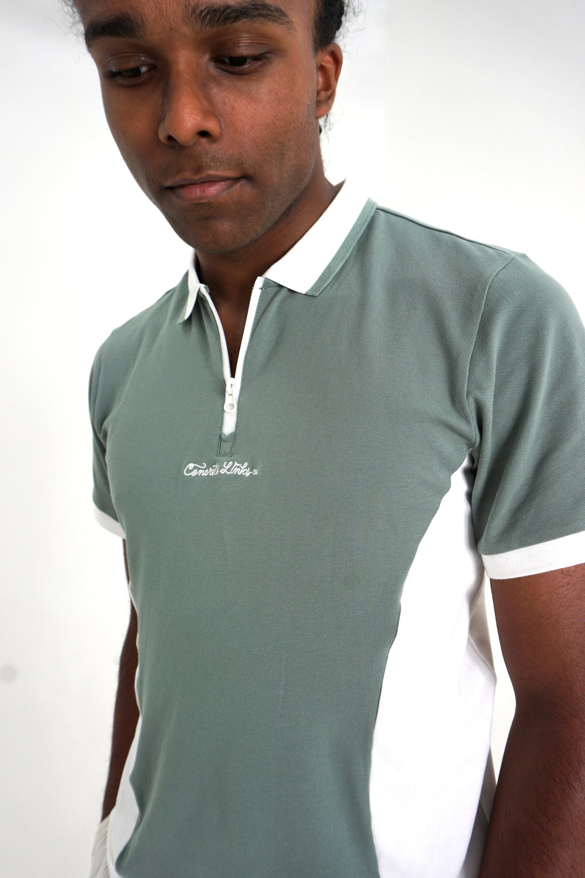 A young man wearing a sage green and white colorblock polo shirt with a white collar and zipper placket. The shirt features a small white script logo on the left chest that reads 'Cruel Kids'.