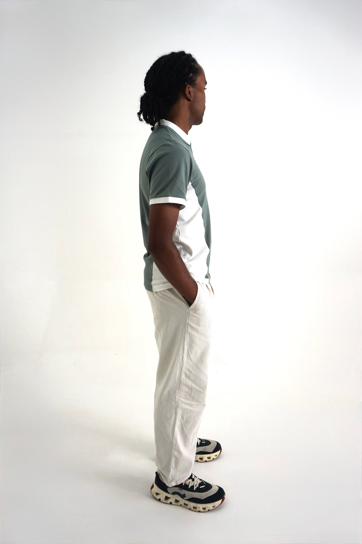 A man standing in profile wearing a short-sleeve polo shirt with a gray and white color-blocked design, white pants, and black and white sneakers, photographed against a plain white studio background.