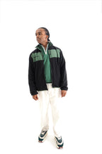 A man modeling a black and green color-blocked zip-up jacket over a green shirt, paired with white pants and green and white sneakers, against a solid white background.