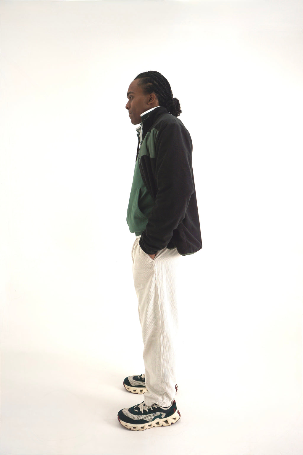 A man standing in profile wearing a black and green color-blocked zip-up jacket, off-white trousers, and dark sneakers with white soles, photographed against a seamless white studio background.