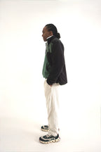 A man standing in profile wearing a black and green color-blocked zip-up jacket, off-white trousers, and dark sneakers with white soles, photographed against a seamless white studio background.