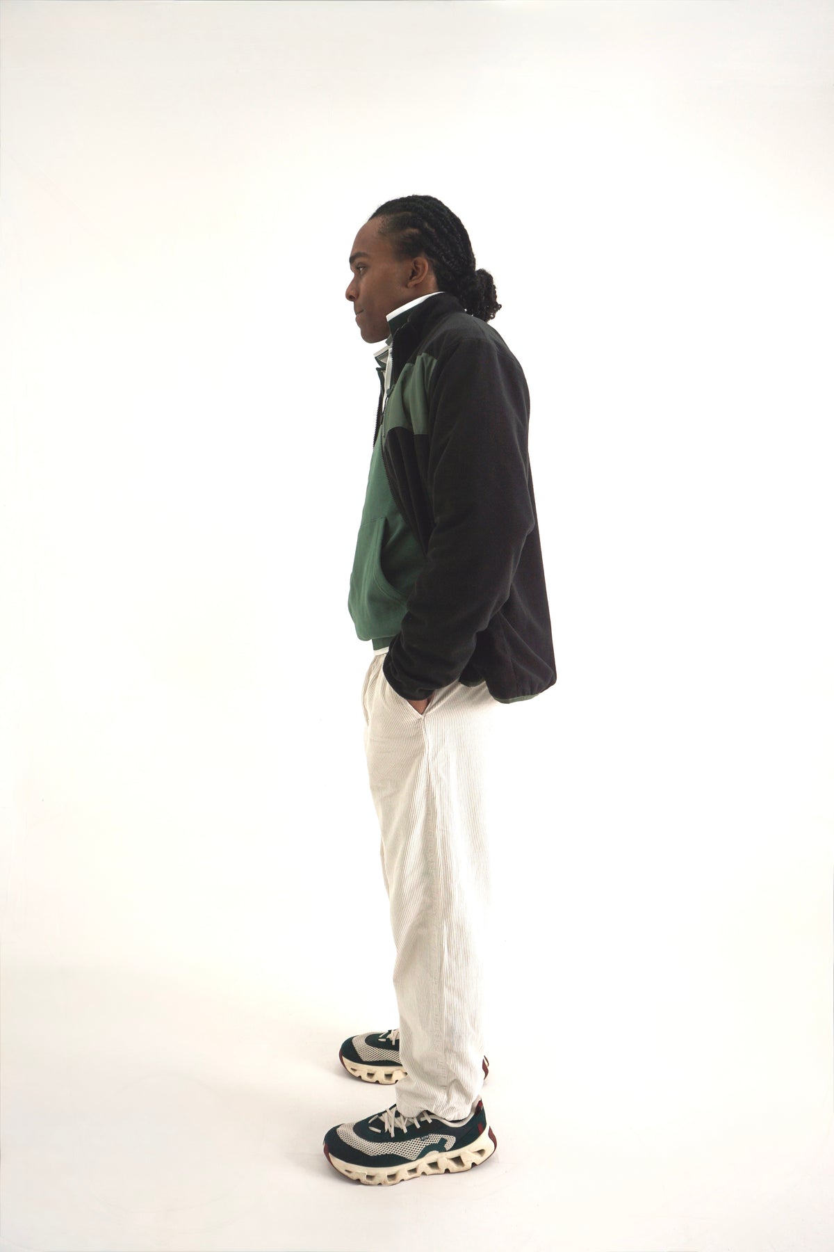 A man standing in profile wearing a black and green color-blocked zip-up jacket, off-white trousers, and dark sneakers with white soles, photographed against a seamless white studio background.