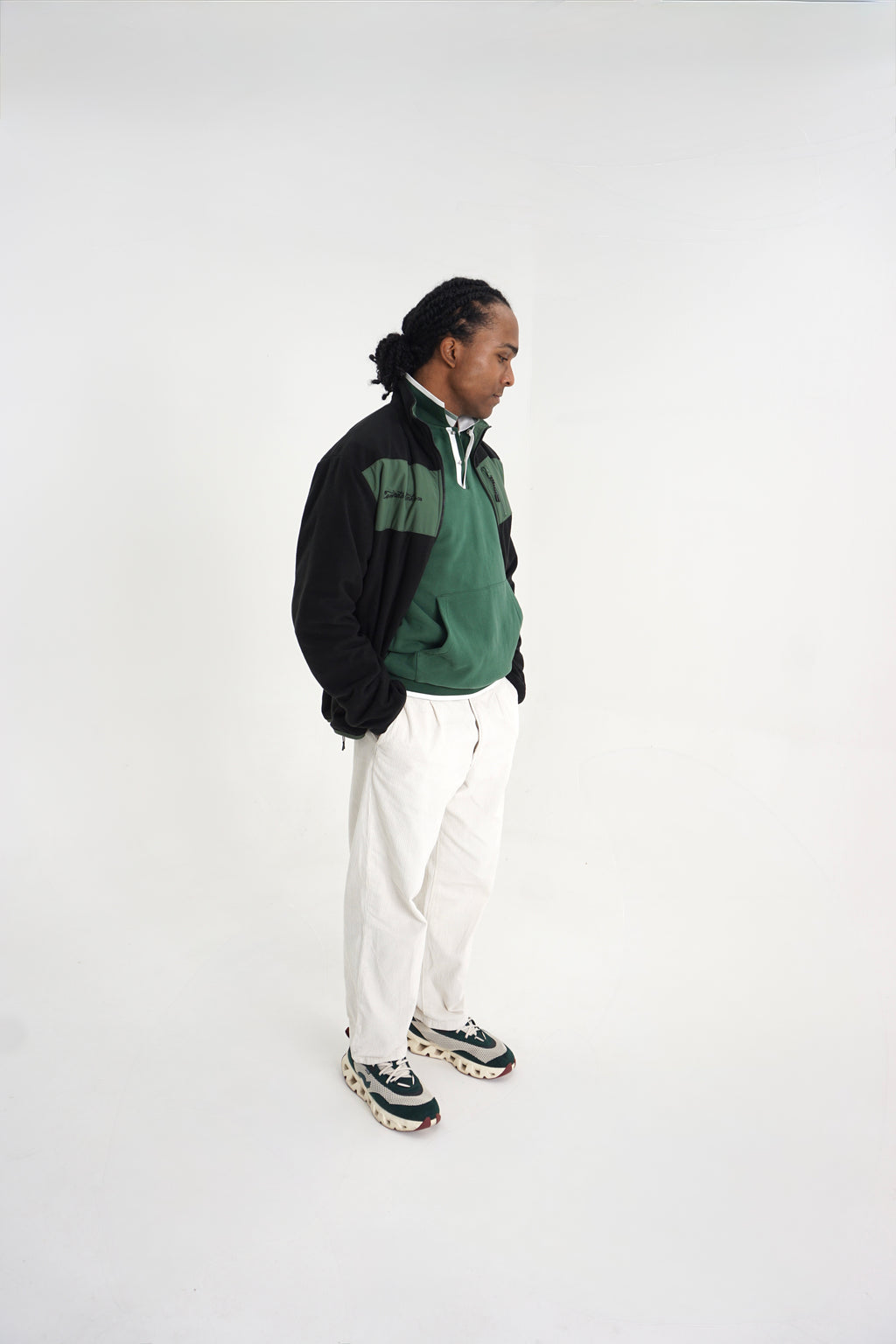 A man stands in profile wearing a green and black quarter-zip pullover, white pants, and patterned sneakers against a plain white studio background.