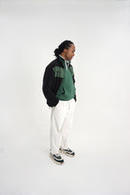 A man stands in profile wearing a green and black quarter-zip pullover, white pants, and patterned sneakers against a plain white studio background.