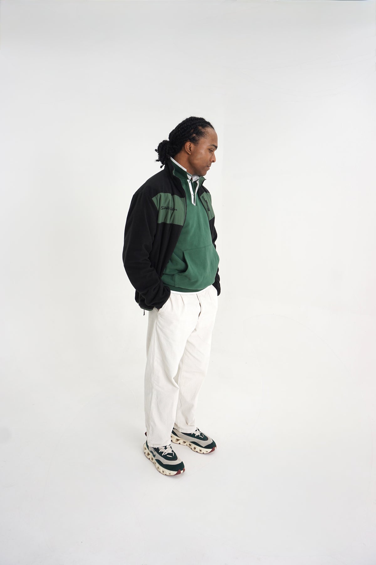 A man stands in profile wearing a green and black quarter-zip pullover, white pants, and patterned sneakers against a plain white studio background.