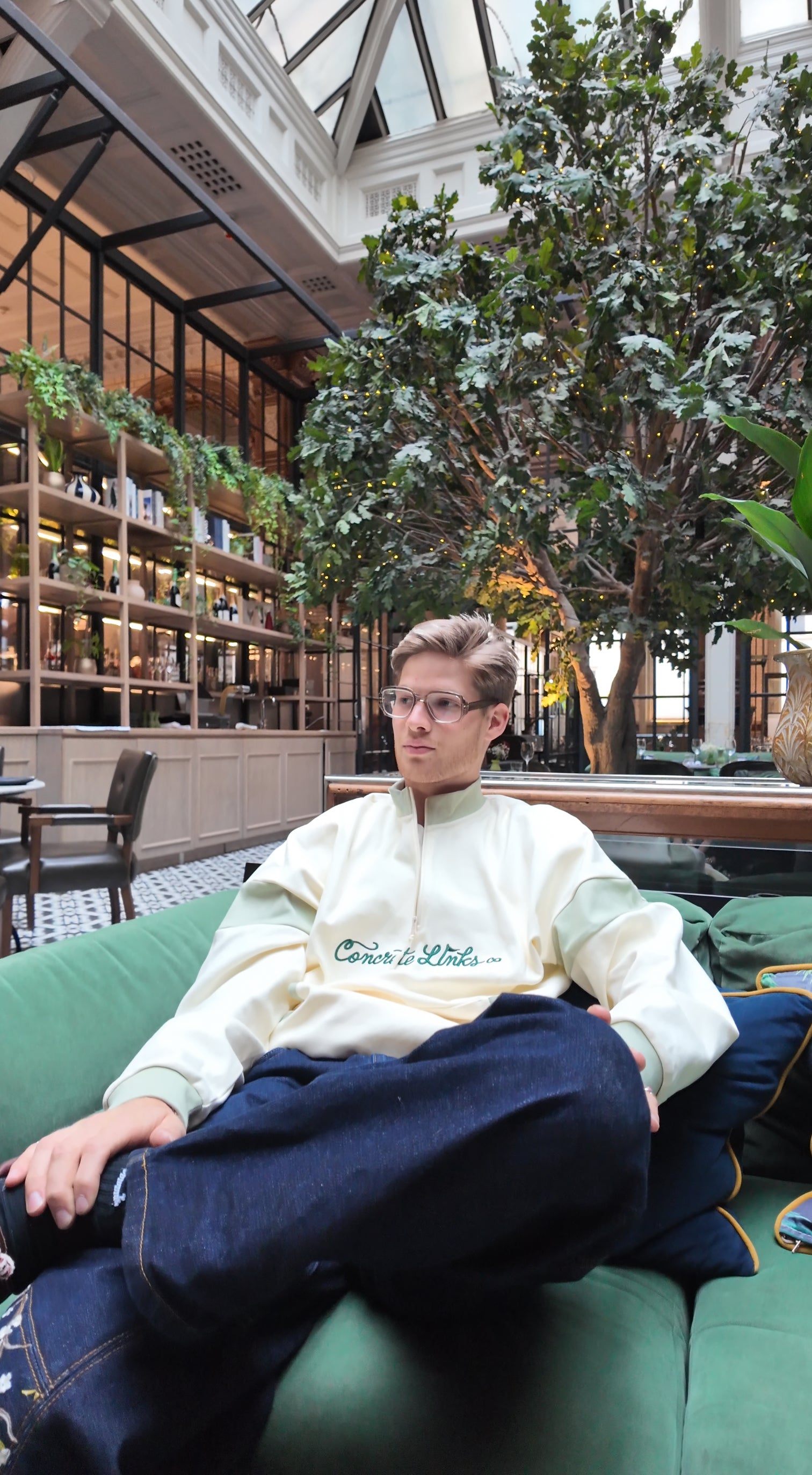 Person sitting on a green couch in a modern indoor setting with plants and bookshelves.