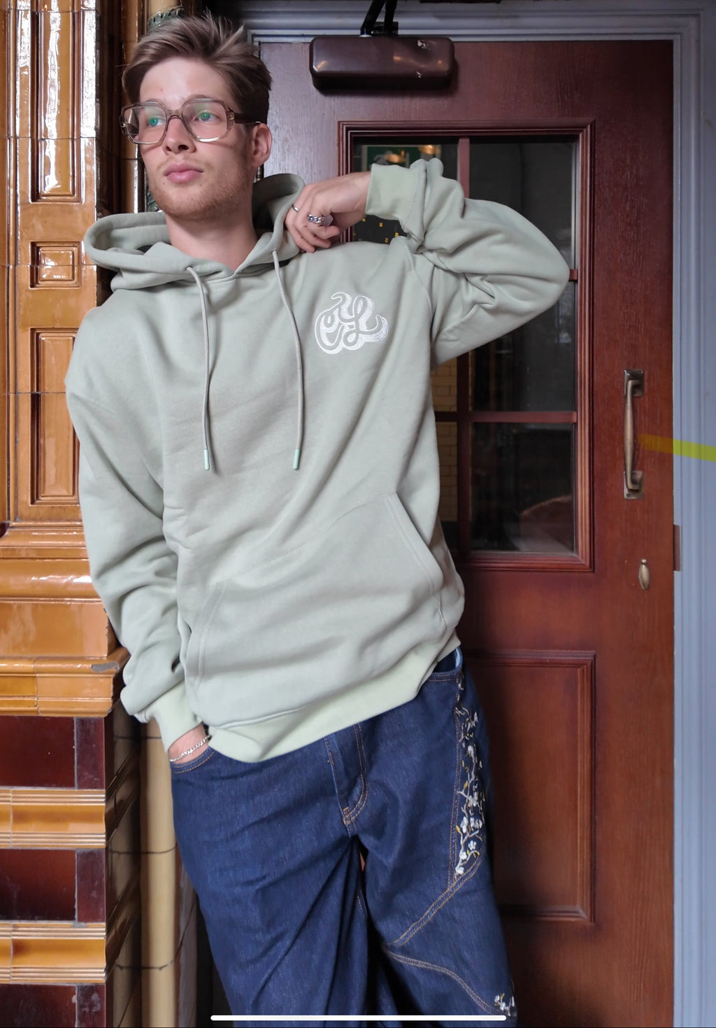 Person wearing a light gray hoodie with a logo, standing in front of a wooden door.
