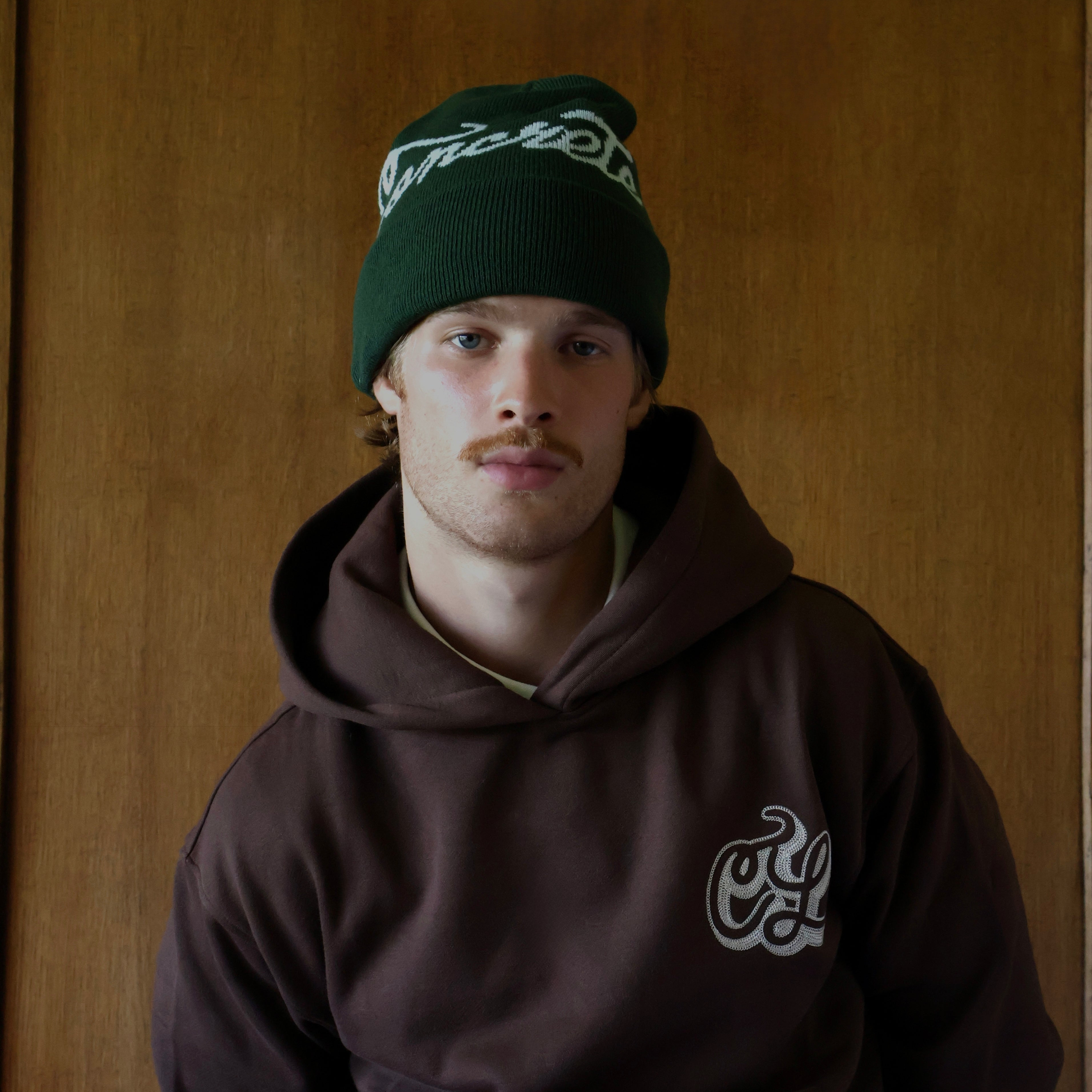 A man wearing a dark brown hoodie with a white graphic on the left chest and a dark green beanie with white script, standing against a wooden door background.